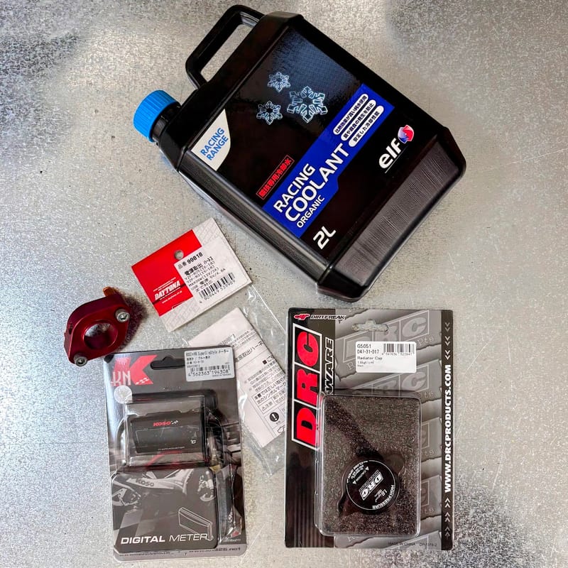 使う部品を並べたところ。elf RACING COOLANT ORGANIC の2L缶、デイトナの電源取出ハーネス 99818、赤いサーモスタットキャンセル、KOSO の水温計、DRC のラジエターキャップ