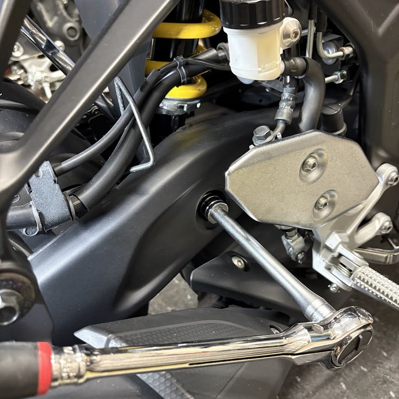 車体右側から、エクステンションバーを継いだラチェットでリヤショックアブソーバーAss'yボルト（下側）を回しているところ